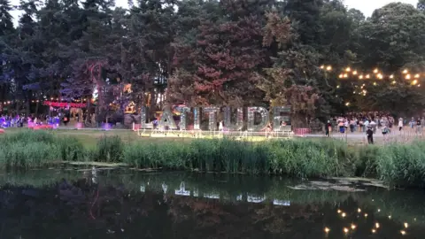 Jon Wright/BBC A big sign saying Latitude on the side of a riverbank. There are light in the trees and woodland behind.