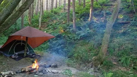 Veterans Response CIC A fire is burning in among a pile of stones on a woodland floor. A tent with a rain sheet hung above it has been pitched nearby. The woodland is thick with ferns growing between the trees.