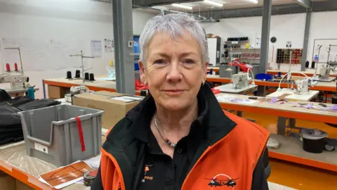 A woman in a bright orange company-branded waistcoat standing in a workshop among benches of sewing machinery and equipment