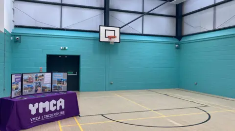 BBC News A sports hall with a basketball hoop and pale blue walls. There is a table covered with a purple cloth. Written on the cloth is "YMCA Lincolnshire", and on the table is a display with photographs