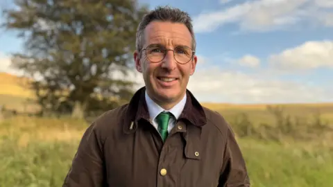 Andrew Muir is wearing a brown coat, blue shirt and green tie. He has silver glasses on. His hair is short and grey. Behind him is open grassland with a single tree. 