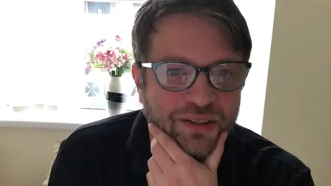 Matty Young, who has floppy brown hair, a beard and moustache and glasses, wearing a black polo shirt and sat with his chin in his hand