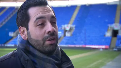 A man wearing a checked scarf and a black coat talking to someone off-camera. He's in a football stadium and there's blue raked seating behind him.