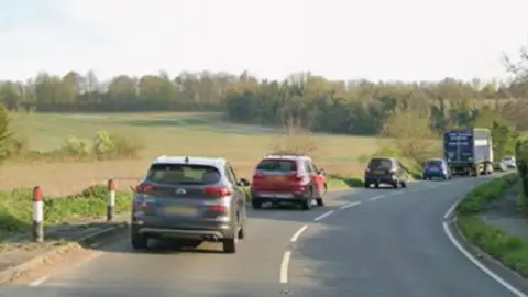 A rural road with a queue of cars on it.