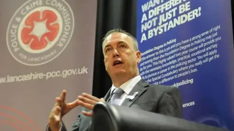 PCC Lancashire police and and crime commissioner Clive Grunshaw with grey hair wearing a grey suit delivers a speech with a police and and crime commissioner poster in the background.