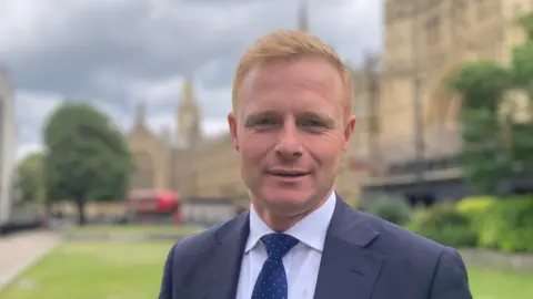 Robbie Moore, who has short ginger hair. He is standing outside the Houses of Parliament and wearing a blue suit with a white shirt and blue tie.