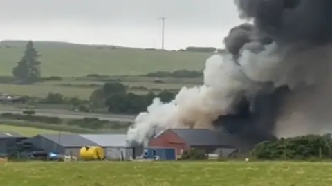Fubar News Large plumes of thick black and grey smoke rise from an industrial site in Tipperty, near Ellon, as firefighters tackle a major blaze. Agricultural and industrial buildings are visible in the foreground, with open countryside stretching into the background. 