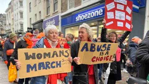 Three women hold three signs as they walk down the high street with the rest of the march. One sign says Womans March, another says abuse is a choice and the third has several pieces of paper and the largest text on it says no means no