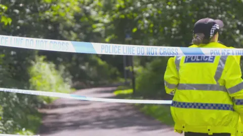 A police officer wearing a hi-viz jacket with her back to the camera. She is standing behind a blue and white strip of police tape.
