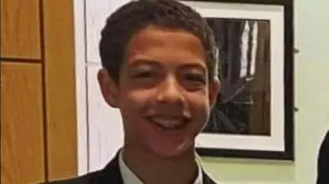 Noah Donohoe, a boy with dark hair, wearing a black blazer, with a white shirt and green, black and white tie.