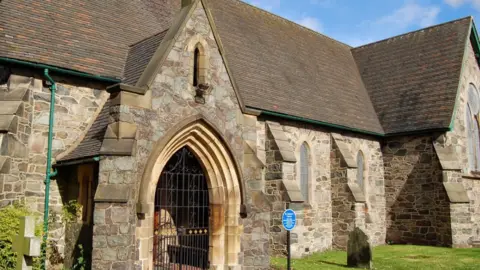 Google The arched entrance to a stone-built church, with a gate across it.  There is a blue heritage plaque to the right of the door 