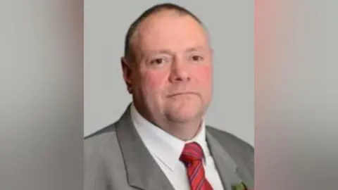 City of Wolverhampton Council A clean shaven man with very short dark hair in a grey suit with a red tie and white shirt is looking at the camer with a slight smile