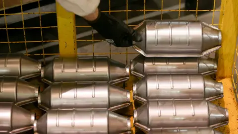 Getty Images Rows of catalytic converters - silver, metallic cylinders - are stored in a yellow cage. A black-gloved hand is reaching down and placing another converter on top of the stack. 