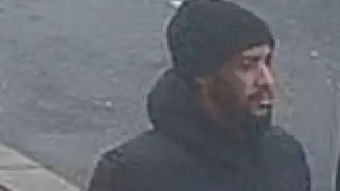 CCTV image of a man with a beard in a black coat and black woolly hat walking down the street