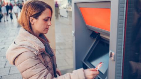 Woman in beige coat with auburn hair about to insert a bank card into an ATM 