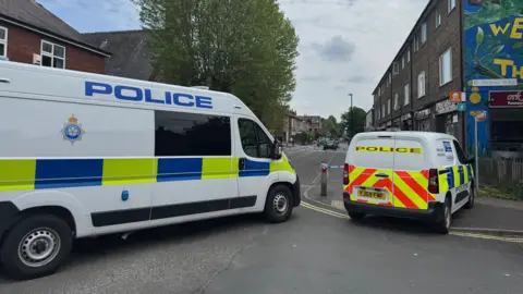 The image shows a cordoned off road with a large white police van on the left and a smaller police vehicle on the right. The rear displays high-visibility red and yellow chevron markings. The word POLICE is printed across the back.
The registration plate reads YJ69 FMF. Between the vehicles, blue-and-white police tape stretches across the road. The street extends into the distance with terraced buildings on both sides.