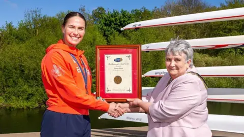 Durham County Council Lauren Irwin and Joan Nicholson are jointly holding a frame with a medal and a certificate inside it. They are standing in front of a lake with rowing boats behind, and a both smiling at the camera and shaking hands.