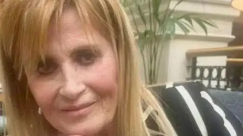 PA Media Jennifer Abbott has straight long blonde hair and is wearing charcoal grey eye make up and a black and white striped top. There is a green pot plant in the background.