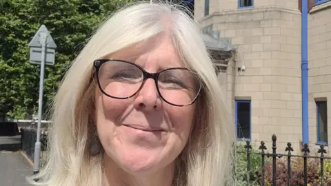 An woman with long bright white hair and glasses smiling at the camera. She stands in front of a grey/brown brick building with the back of a road sign near to her. 