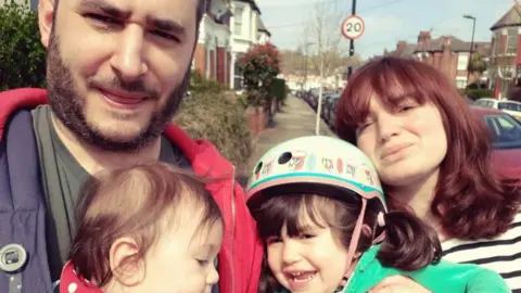 A family photo showing Hannah and her husband, and they are holding their two young children, in a selfie style photo on an English street.