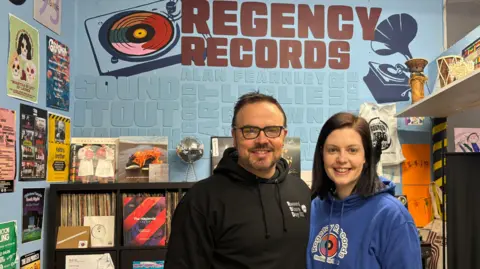 BBC/Eve Connor Stephen Thompson and his partner Rebecca. They are standing side-by-side in front of a sign that says Regency Records. Stephen is wearing a black hoodie with a small logo on the left side of the chest that says Record Store Day, and Rebecca is wearing a blue hoodie that has a logo in the centre saying Regency Records. Stephen has black hair, and is wearing glasses. Rebecca has dark brown hair.