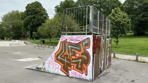 A skatepark ramp is in the centre of the image. It is steep with metal railings on top. It is covered in coloured grafitti. The skatepark surrounds it in the foreground. In the background is the green grass of St George Park and a number of trees.