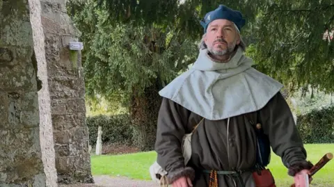 Stuart Huntley A man dressed in medieval attire walking past a church building with grass and graves seen behind. A large tree is also covering the skyline.