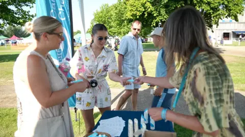 Shaun Whitmore/BBC A lady giving out free suncream at a stall to two ladies both dressed in white and each holding fans.