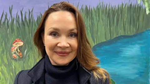 Lorna Hackett - a woman with brown hair smiling at the camera. Behind her is a colourful picture of water with pond animals.