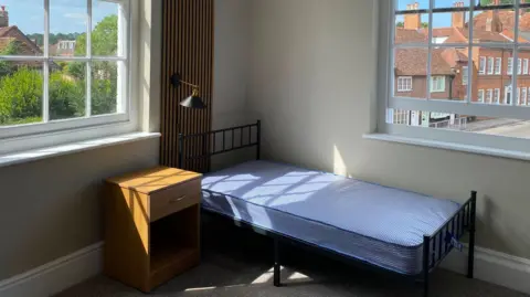 Alabaré A bedroom in the Alabaré Veterans House in Salisbury featuring a black metal-framed single bed with a blue stripey mattress, a wooden bed-side table and a black wall lamp, set against beige walls and two large windows with the street, houses and trees outside. 