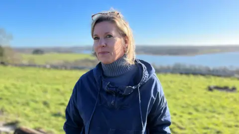 Amber Lort-Phillips. Founder of The Big Retreat Festival, wears a blue jumper and coat in a field. She has short blonde hair.