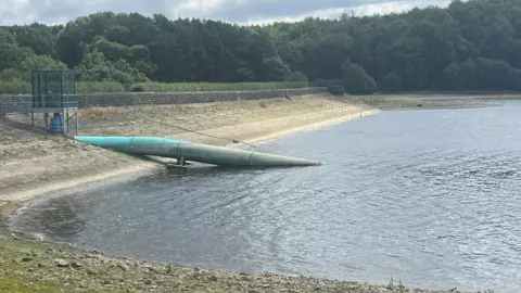 A dried out reservoir in Leeds. A large turquoise pipe can be seen going into the water. There are trees in the background.