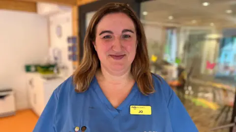 A woman in blue medical scrubs smiles at the camera. She has straight brown hair and a yellow name badge that says "Jo". There is a kitchen behind her.