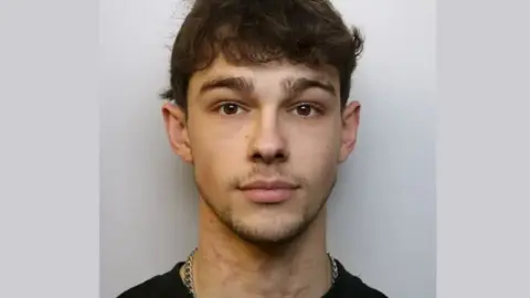 Mathew McGrath looking straight into the camera in his mugshot photo. He has curly brown hair, thick eyebrows and a thin beard and moustache. He wears a black t-shirt and a thick silver chain. 