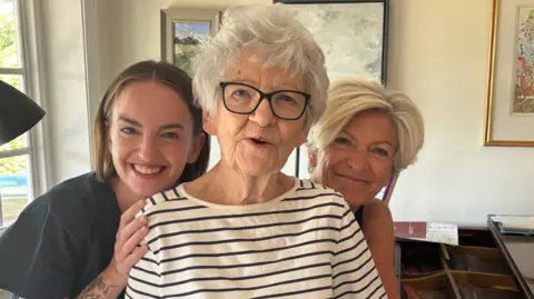 Luke Deal/BBC A young woman with short brown hair and a middle-aged woman with short blonde hair stand behind an elderly woman, with their hands on her shoulders, as they smile at the camera. The elderly woman has short grey hair, glasses and is wearing a white long-sleeved top with black stripes. 