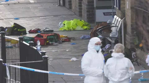 PA Media Two emergency service workers in forensic suits stand in the right hand corner of the image. Police cordon has segregated an area where paramedic equipment, clothing and other belongings lie on the floor