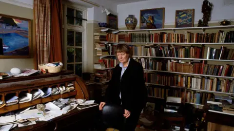 Sophie Bassouls/Sygma/Sygma via Getty Images Iris Murdoch standing in a room at her home surrounded by books, her desk covered in papers and artwork such as paintings. 