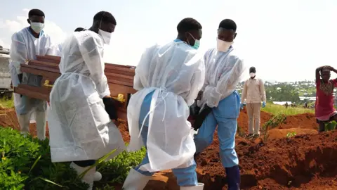 Volunteers in blue scrubs, rubber boots and white gowns carry a coffin towards a dug grave. A few people, including one holding a spade, watch on from the side.