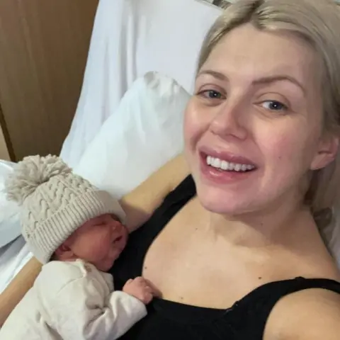 Julie Keegan Blonde woman smiling in a hospital bed while holding newborn baby. She is wearing a black tank top. Baby is wearing a white babygrow with a white wooly hat.