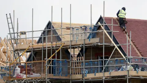 A generic image of a building site, with a builder in a high-vis jacket perched on the roof of a house. Scaffolding surrounds two properties, with a third in the background on the left