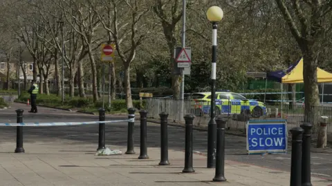 Blue police tape is attached around black bollards, with a police car seen in the background.