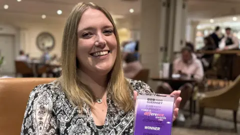 BBC A young woman with shoulder length blonde hair with a middle parting, smiles at the camera. Sitting in a leather chair in a bar, wearing a black and white paisley top, holding a purple and white rectangular award in her hands.