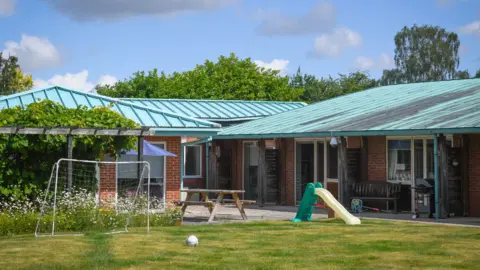 Acorns A one-storey building with green-coloured roofing on a sunny day. Trees are visible behind the building and in front is a grassy area with a football, a goal, a small slide and a picnic bench.