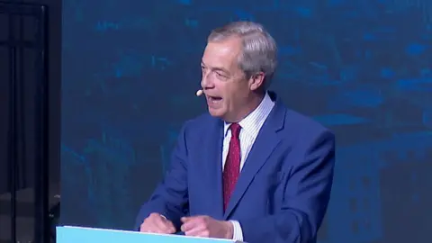 Nigel Farage stands at a powder blue podium and is speaking on stage in a blue suit and red tie. There is a blue backdrop behind him.