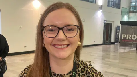 A close up image of a girl, smiling at the camera. She has light brown hair with a side parting. She is wearing gold earrings and a leopard print top. 