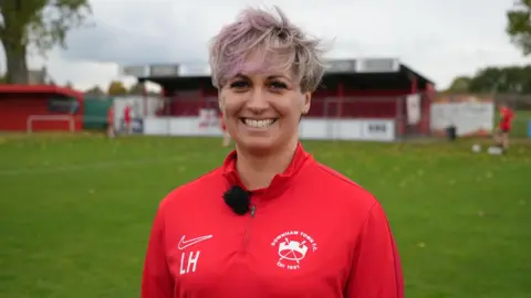 Shaun Whitmore/BBC Woman with light pink hair is smiling. She is wearing a red football shirt with the logo Downham Town FC in white.