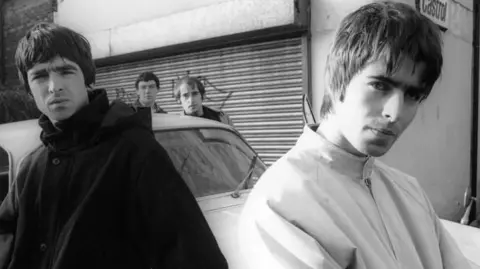 Black-and-white photo of Oasis in 1993. Liam Gallagher stands in the foreground with arms crossed, while Noel leans against a vintage white Jaguar XJ6 behind him. The rest of the band are posed around the vehicle on a backstreet.