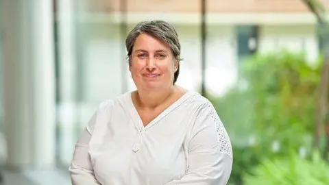 University of Aston A woman with short dark grey hair smiles at the camera. She wears a white v-neck top. The background is blurred but shows large windows and green plants.