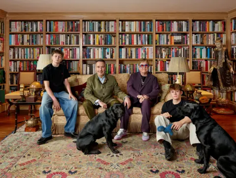 National Portrait Gallery/Catherine Opie, Regen Projects and Thomas Dane Gallery Catherine Opie's photo of Elton and his family - Zachary, David, Elton and Elijah with the two pet black labradors in the foreground. They are seated on a sofa and rug in a library lined with bookshelves.