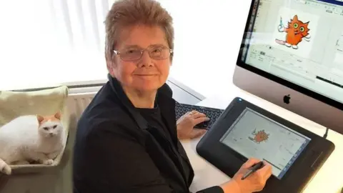Sheila Graber smiling into the camera while working on an animation. She has silver, rectangular glasses and is wearing a black jacket. On her tablet in front of her there is cartoon of an orange cat holding a paintbrush, which is also shown on a larger screen on the desk. Behind her is a white and ginger cat lying down.